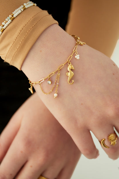 Close-up of gold charm bracelet on wrist, showing seahorse and pearl charms with layered chains