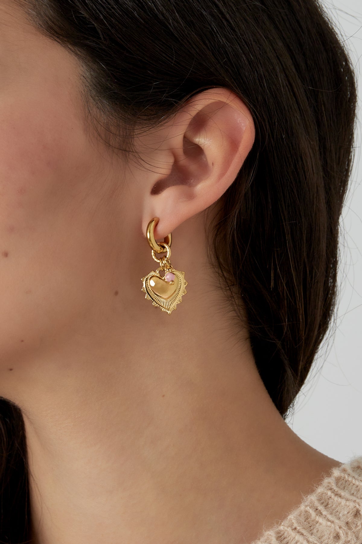 Close-up of a woman's ear wearing gold heart charm earrings with pink stone beads