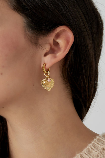 Close-up of a woman's ear wearing gold heart charm earrings with pink stone beads