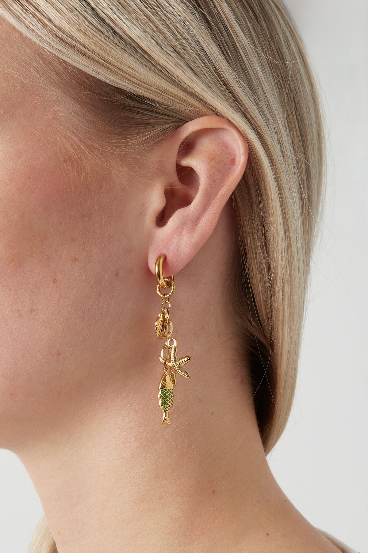 Close-up of woman wearing gold-tone sea charm drop earring with starfish, seashell and green enamel fish