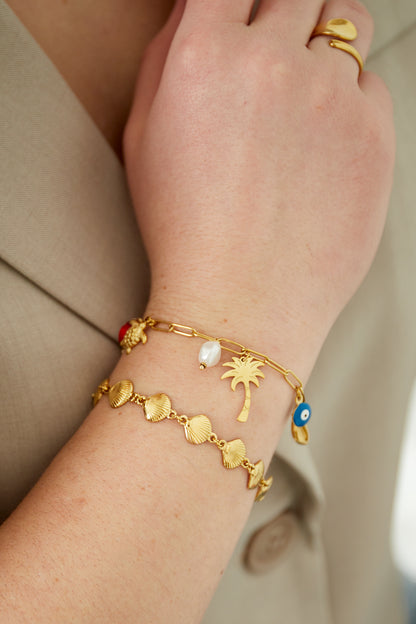 Wrist wearing gold seashell charm bracelet paired with a gold pine tree and pearl charm chain bracelet and a gold ring.