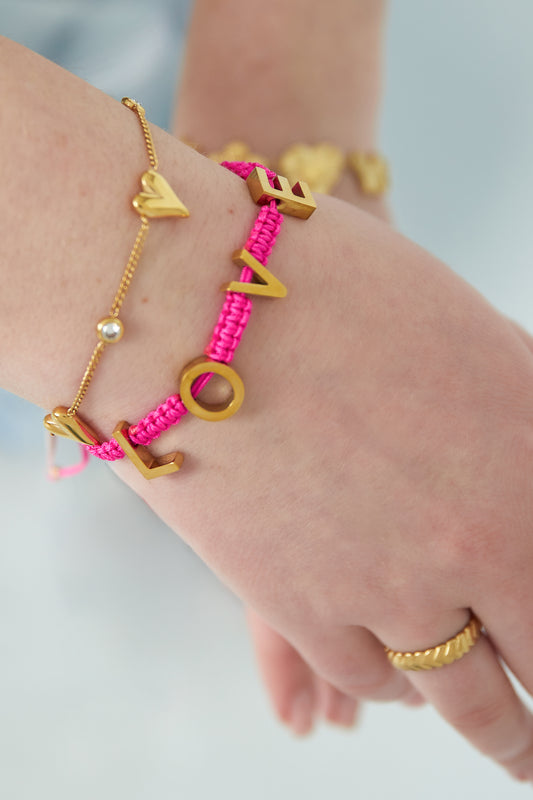Model's wrist with layered bracelets including the fuchsia pink LOVE braided bracelet and gold heart charm bracelet
