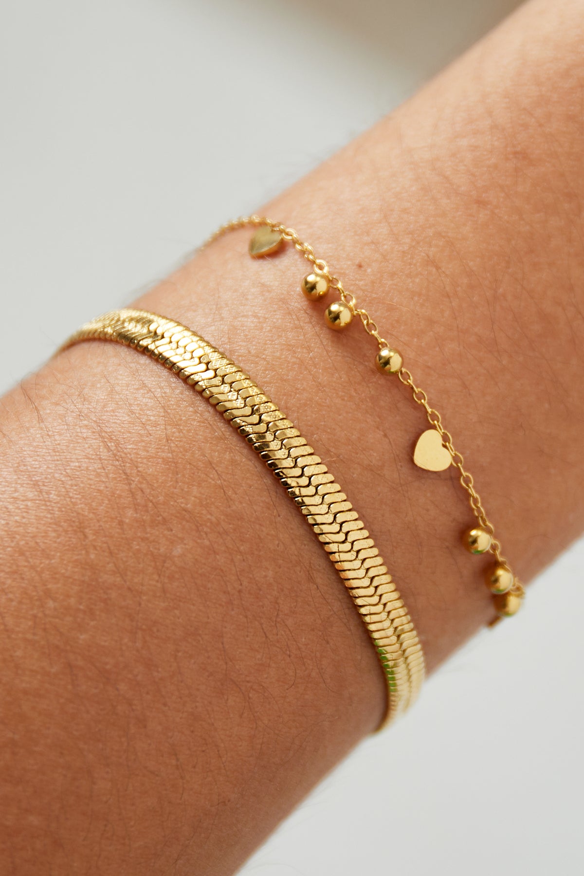 Two gold bracelets stacked on wrist, one snake chain and one delicate chain with heart and bead charms