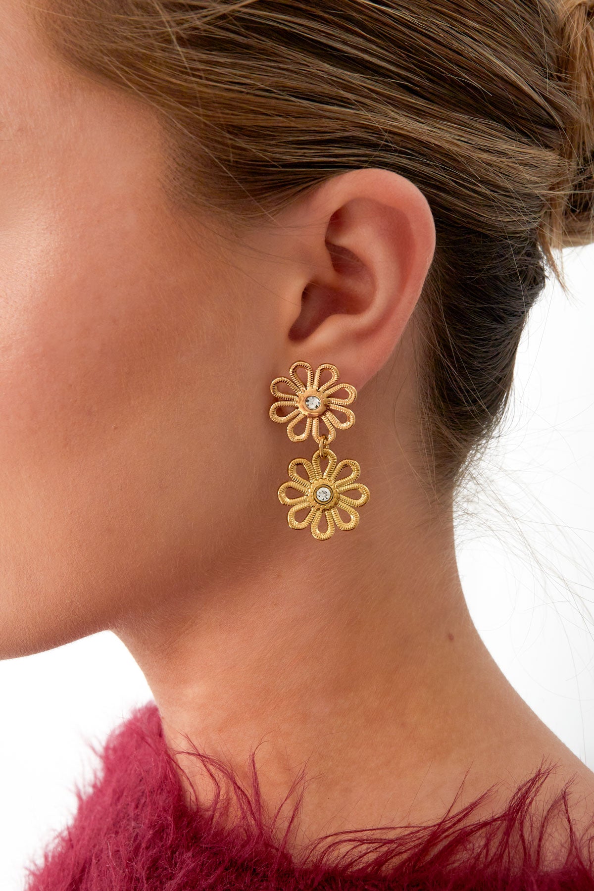 Side profile of a woman wearing gold flower drop earrings with two linked flowers and crystal centers, paired with a fuzzy magenta top