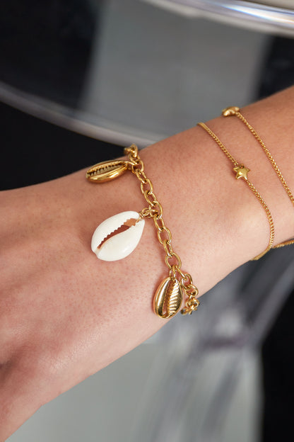 Bracelet worn on a wrist alongside delicate gold chains, showing gold-tone and white shell charms swinging freely
