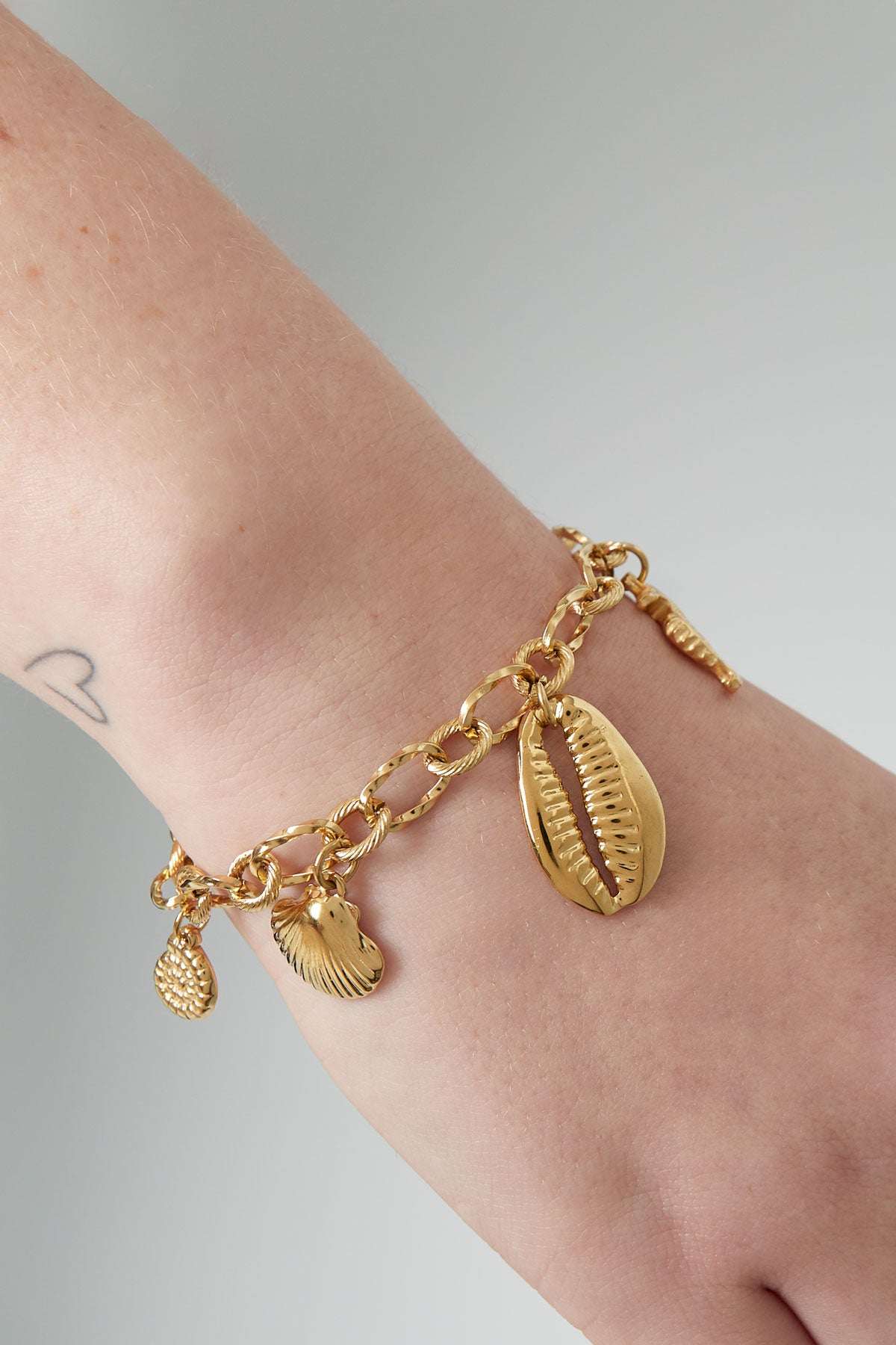 Close-up of gold sea charm bracelet worn on wrist featuring starfish, seahorse, shell, and cowrie shell charms with a small wrist tattoo visible.