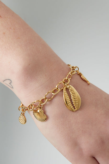 Close-up of gold sea charm bracelet worn on wrist featuring starfish, seahorse, shell, and cowrie shell charms with a small wrist tattoo visible.