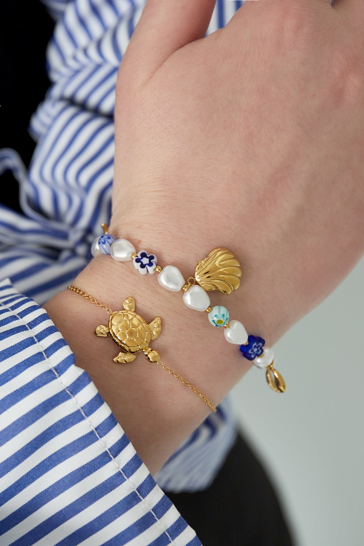 Wrist wearing two bracelets: a gold turtle charm bracelet paired with a colourful bead bracelet with gold shell and floral charms