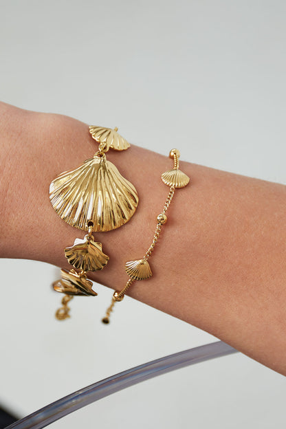 Close-up view of two gold seashell bracelets worn on wrist, showing delicate shell charms and fine chain links