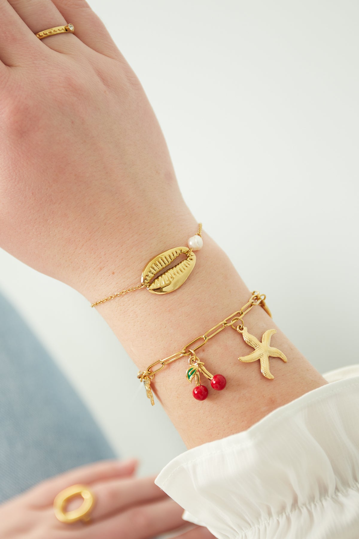 Bracelet with gold shell charm and pearl detail worn on a wrist alongside a charm bracelet with cherries and starfish