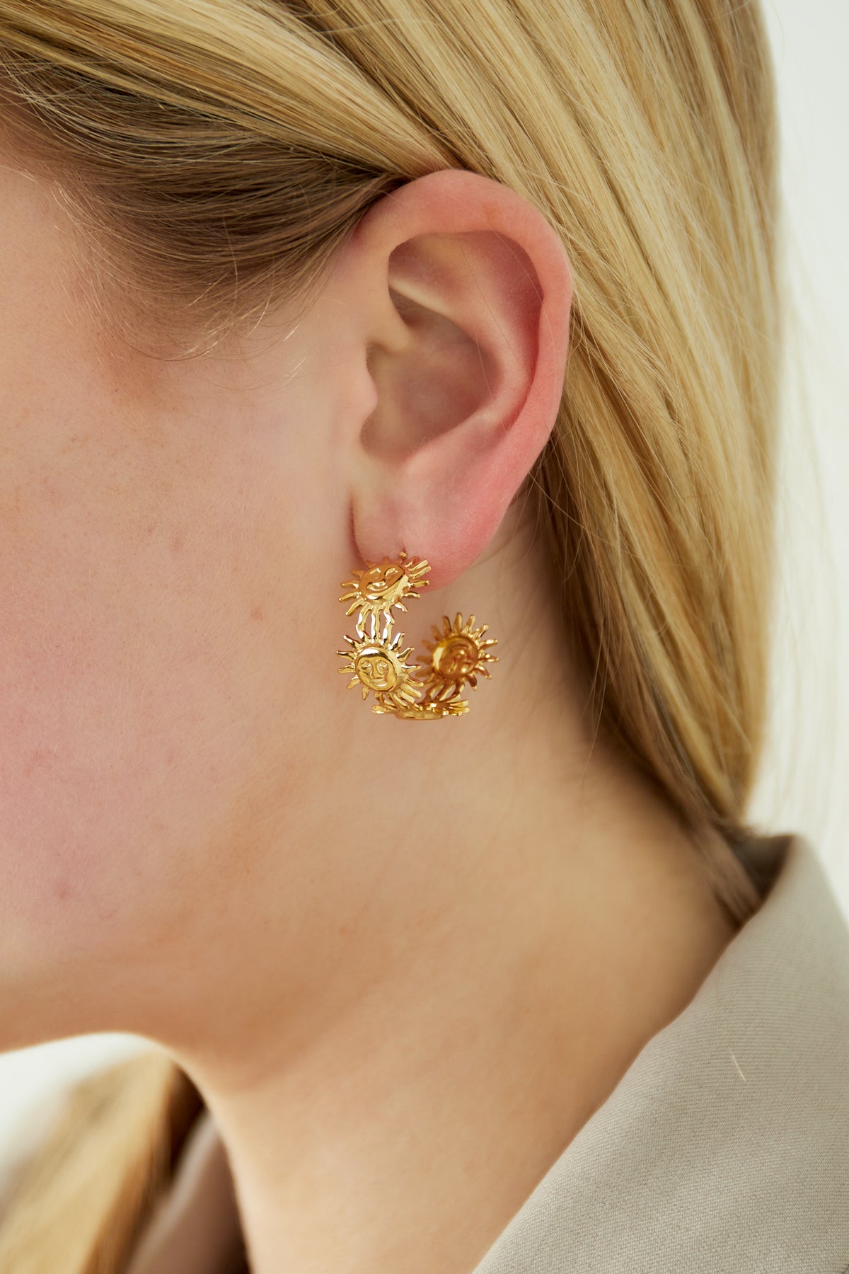 Close-up of a woman's ear wearing gold sun face hoop earring, styled with blonde hair and beige shirt