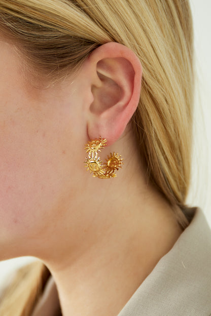 Close-up of a woman's ear wearing gold sun face hoop earring, styled with blonde hair and beige shirt