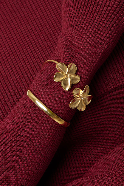 Model wearing gold floral cuff bracelet over burgundy ribbed sleeve next to a smooth gold bangle