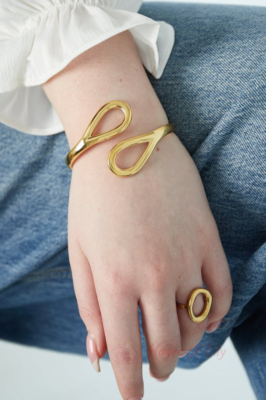 Close-up of a woman wearing a gold-toned cuff bracelet and matching ring with teardrop loops, styled with white blouse and denim jeans