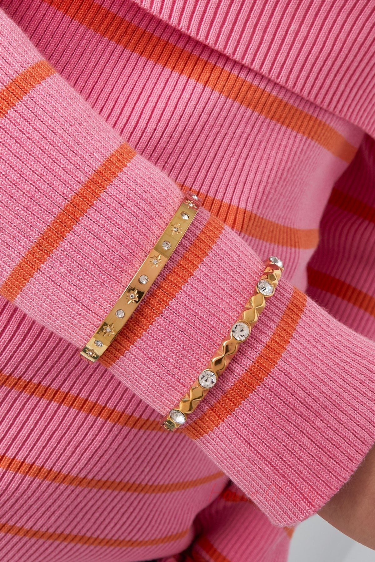 Two gold tone bracelets worn together on wrist with pink and orange striped sleeve - one crystal studded relief bangle and one star-studded bangle
