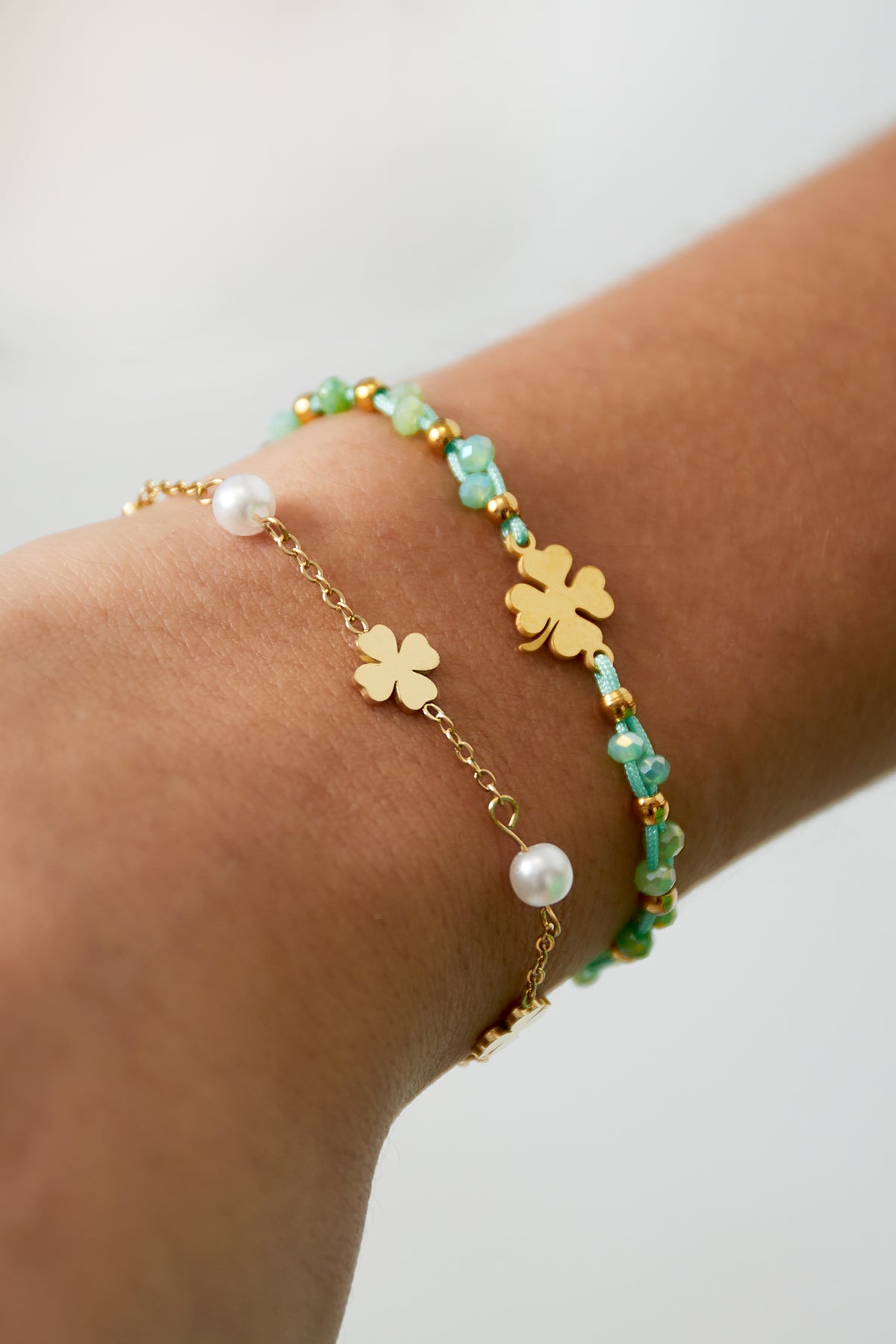 Close-up of a wrist wearing two bracelets: one gold with clover and pearl charms, another with green and gold beads