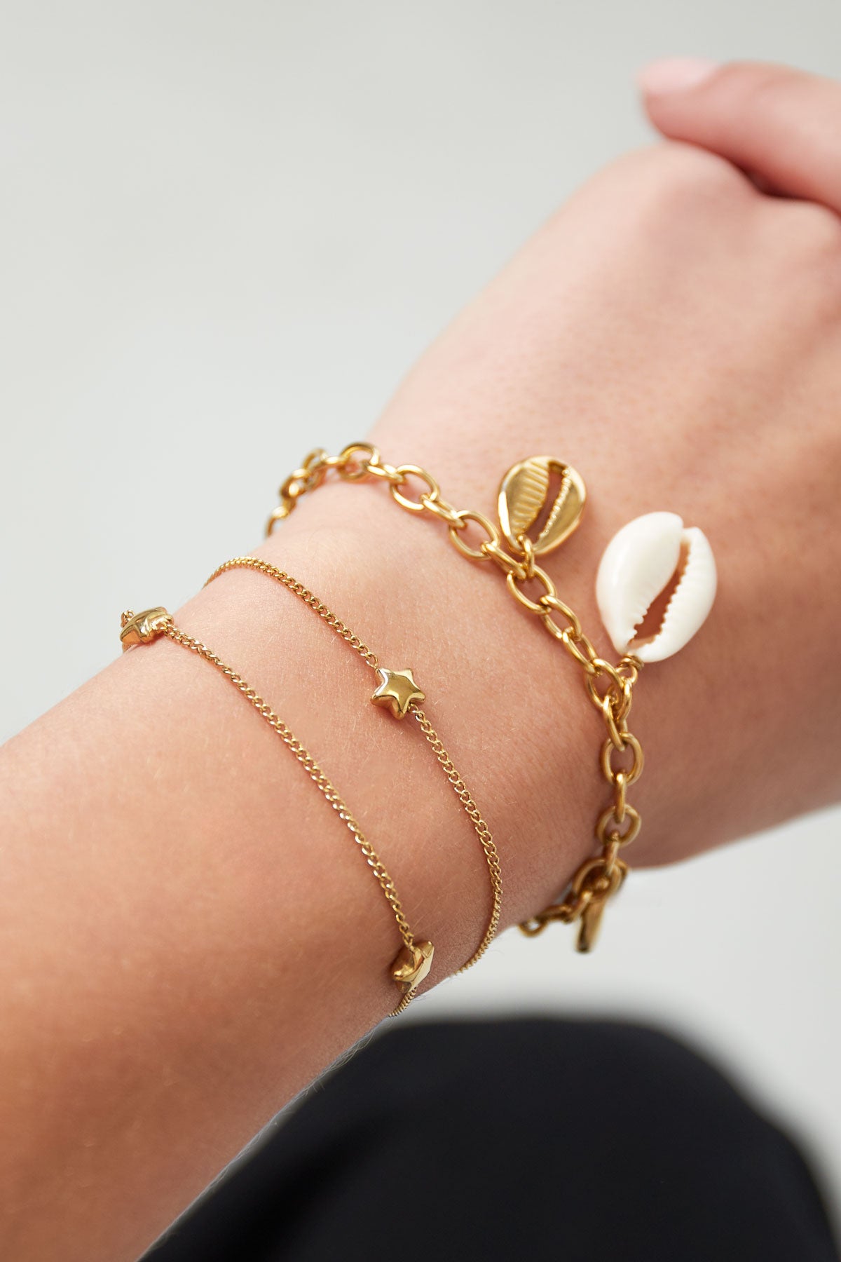 Close-up of wrist wearing two bracelets: a delicate gold star charm bracelet and a chunky gold chain bracelet with shell charms