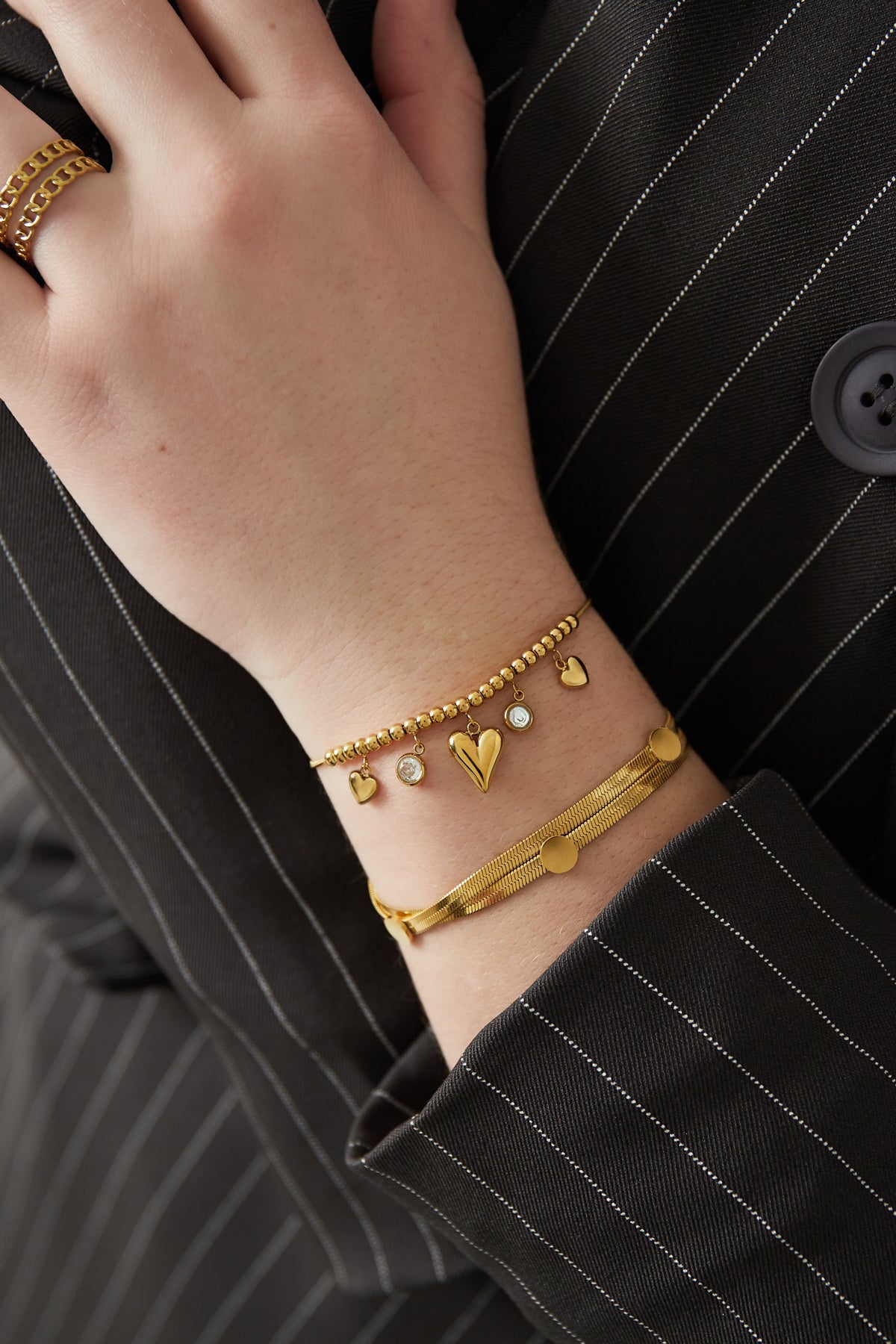 Gold-tone flat chain bracelet with dot accents worn on wrist alongside charm bracelet with hearts and crystals