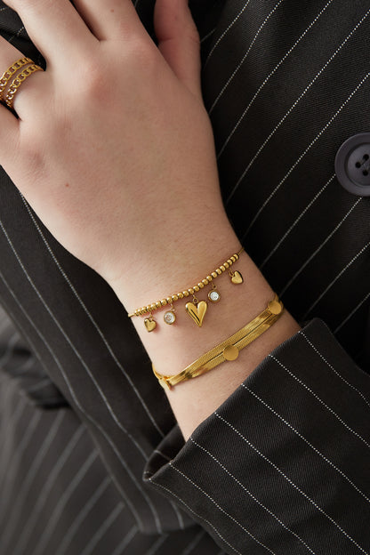 Gold-tone flat chain bracelet with dot accents worn on wrist alongside charm bracelet with hearts and crystals
