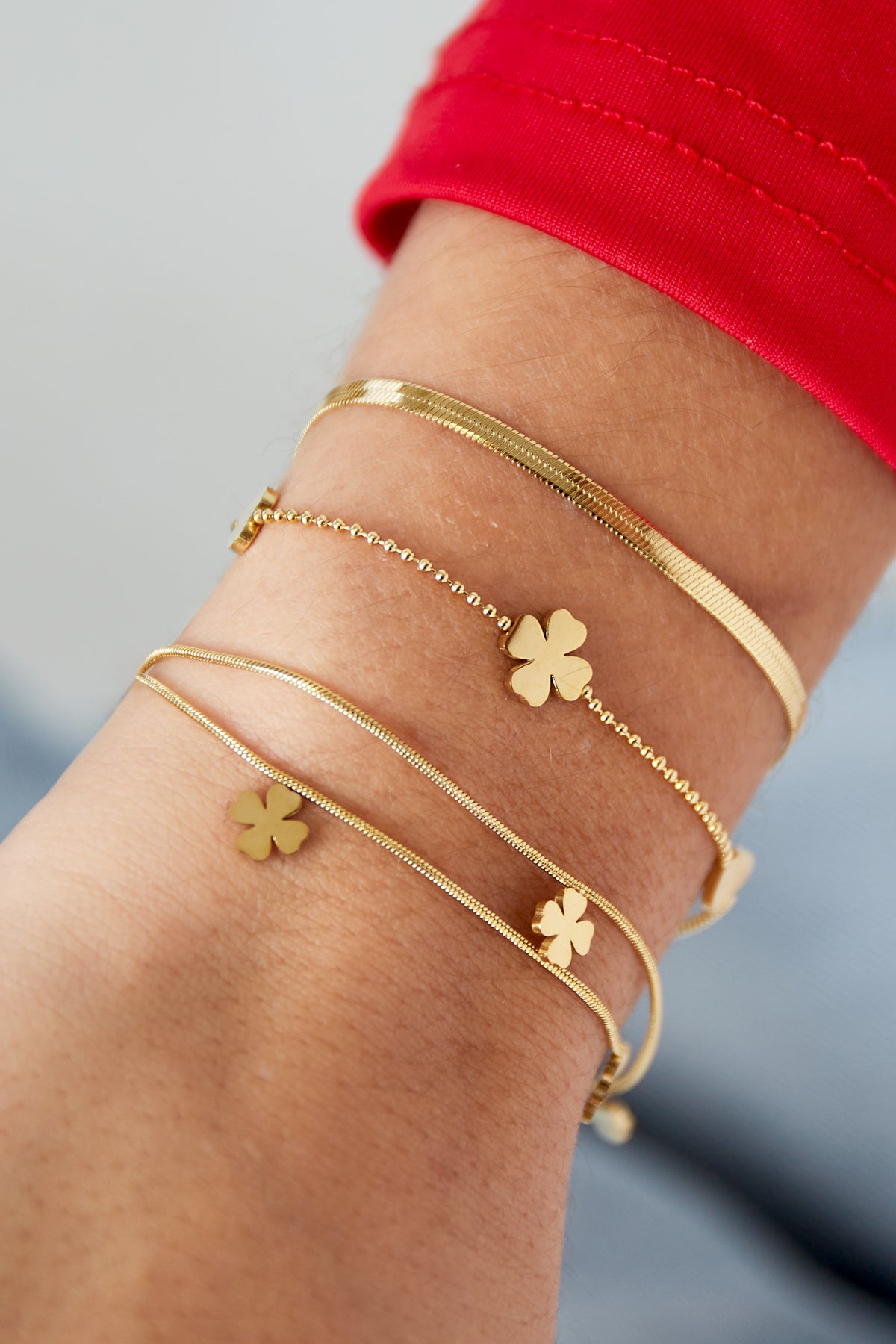 Gold layered charm bracelet with heart and clover charms worn on wrist with red sleeve in background