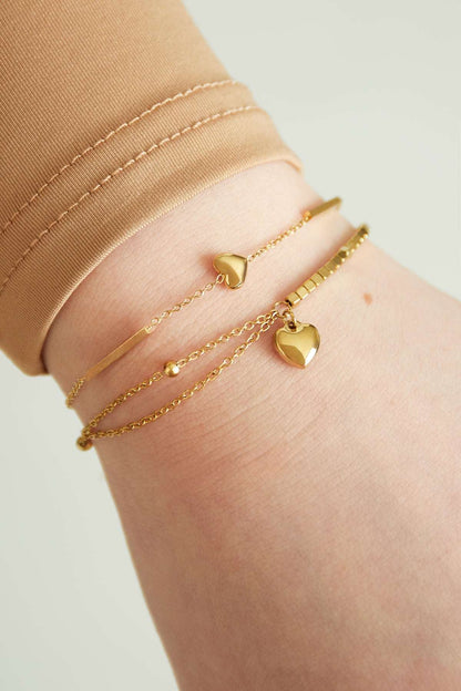 Close-up of gold heart charm bracelet layered with another delicate gold bracelet on wrist