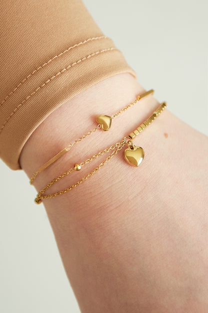 Close-up of gold heart charm bracelet layered with another delicate gold bracelet on wrist
