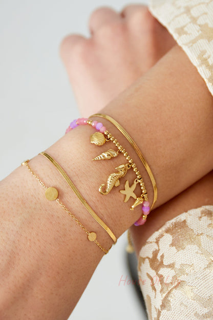Wrist adorned with multiple bracelets including gold minimal coin charm bracelet paired with pink bead and sea-themed charm bracelets