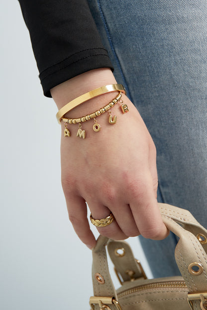 Gold-tone AMOUR charm bracelet worn on wrist alongside gold bangle and ring with blue jeans and beige handbag in background