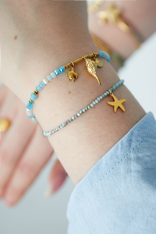 Beaded bracelets worn stacked on a wrist, showing a starfish charm on a blue faceted bead bracelet paired with another gold charm bracelet.