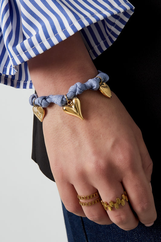 Model wearing blue braided heart charm bracelet with three gold heart pendants on wrist and gold rings