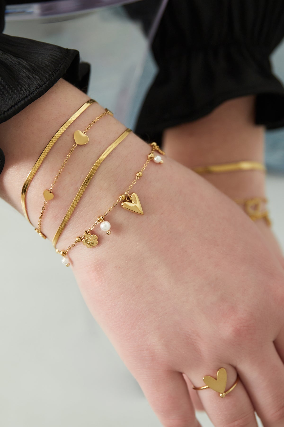 Gold charm bracelet worn on wrist alongside slim gold bands and matching heart ring with black sleeve background