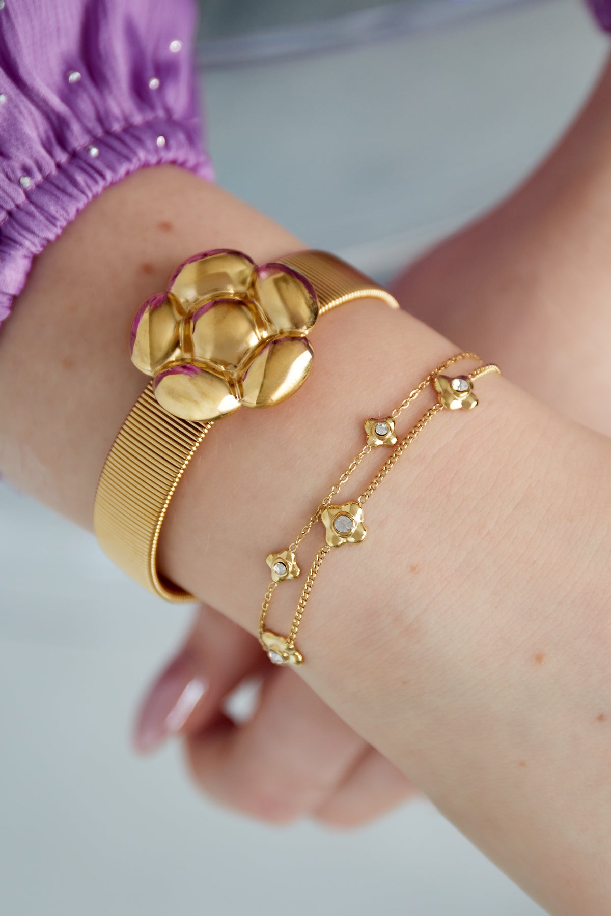 Close up of a wrist wearing gold double chain flower bracelet paired with a wider gold bracelet and a purple sleeve