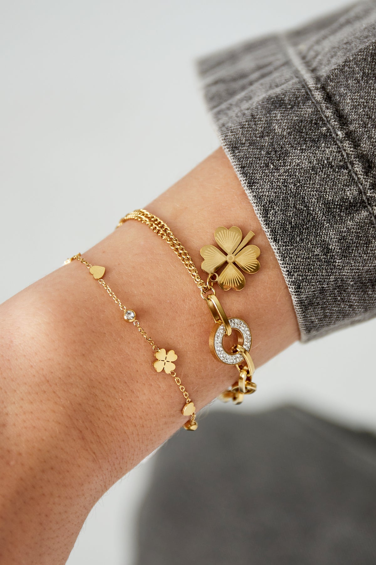 Close-up of wrist wearing two gold bracelets: one charm bracelet with star, heart and clover charms, paired with a thicker link bracelet with clover charm