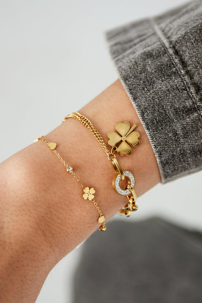 Close-up of wrist wearing two gold bracelets: one charm bracelet with star, heart and clover charms, paired with a thicker link bracelet with clover charm