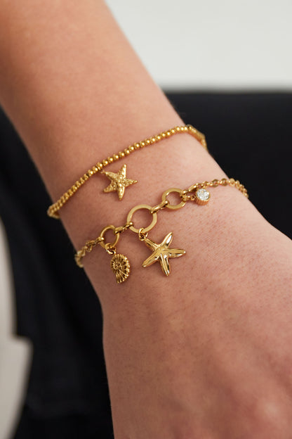 Close-up of wrist wearing two gold-tone bracelets, including starfish charm bracelet with shell and rhinestone details