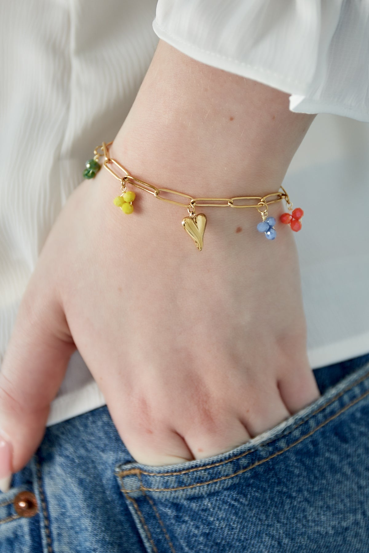 Model wearing gold charm bracelet featuring colourful glass beads and heart charm, styled with denim and white top