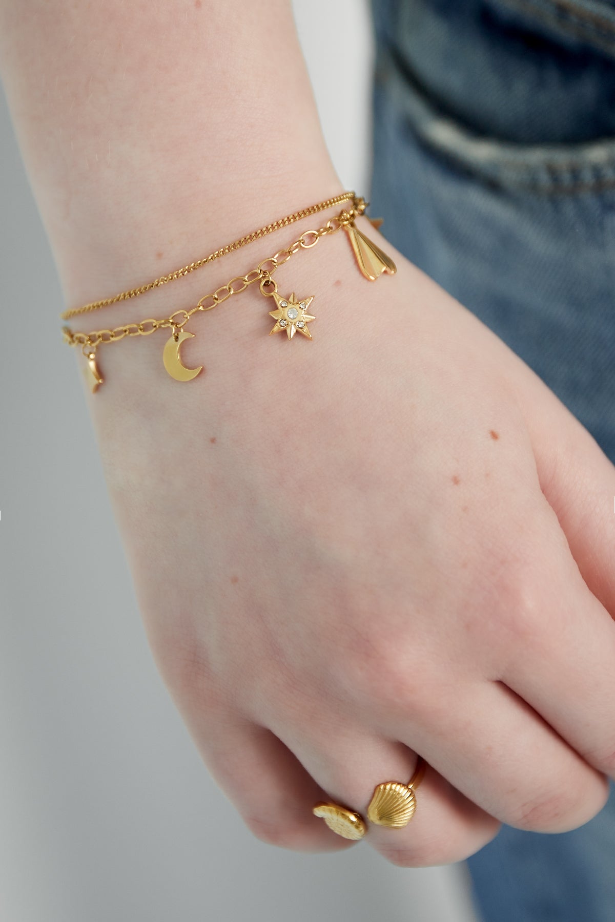 Close-up of the gold-tone charm bracelet worn on wrist paired with a gold shell ring against a casual denim outfit