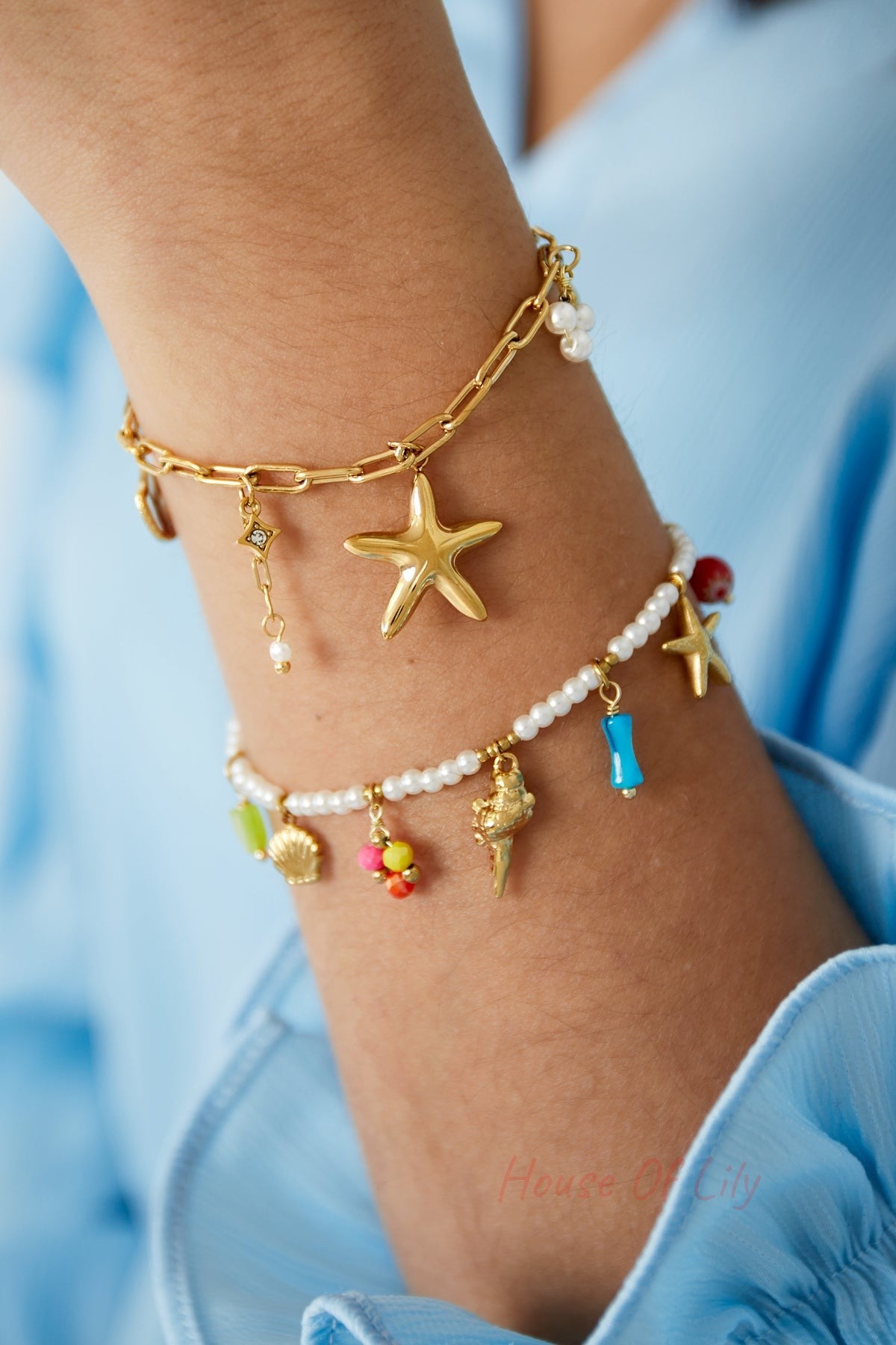 Close-up of two stacked charm bracelets on wrist including gold-tone seaside charm bracelet with starfish and pearl details and colourful beaded bracelet