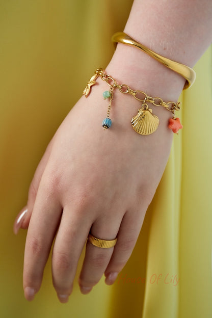 Woman's wrist wearing the gold charm bracelet alongside a smooth gold bangle, showing playful marine-inspired charms
