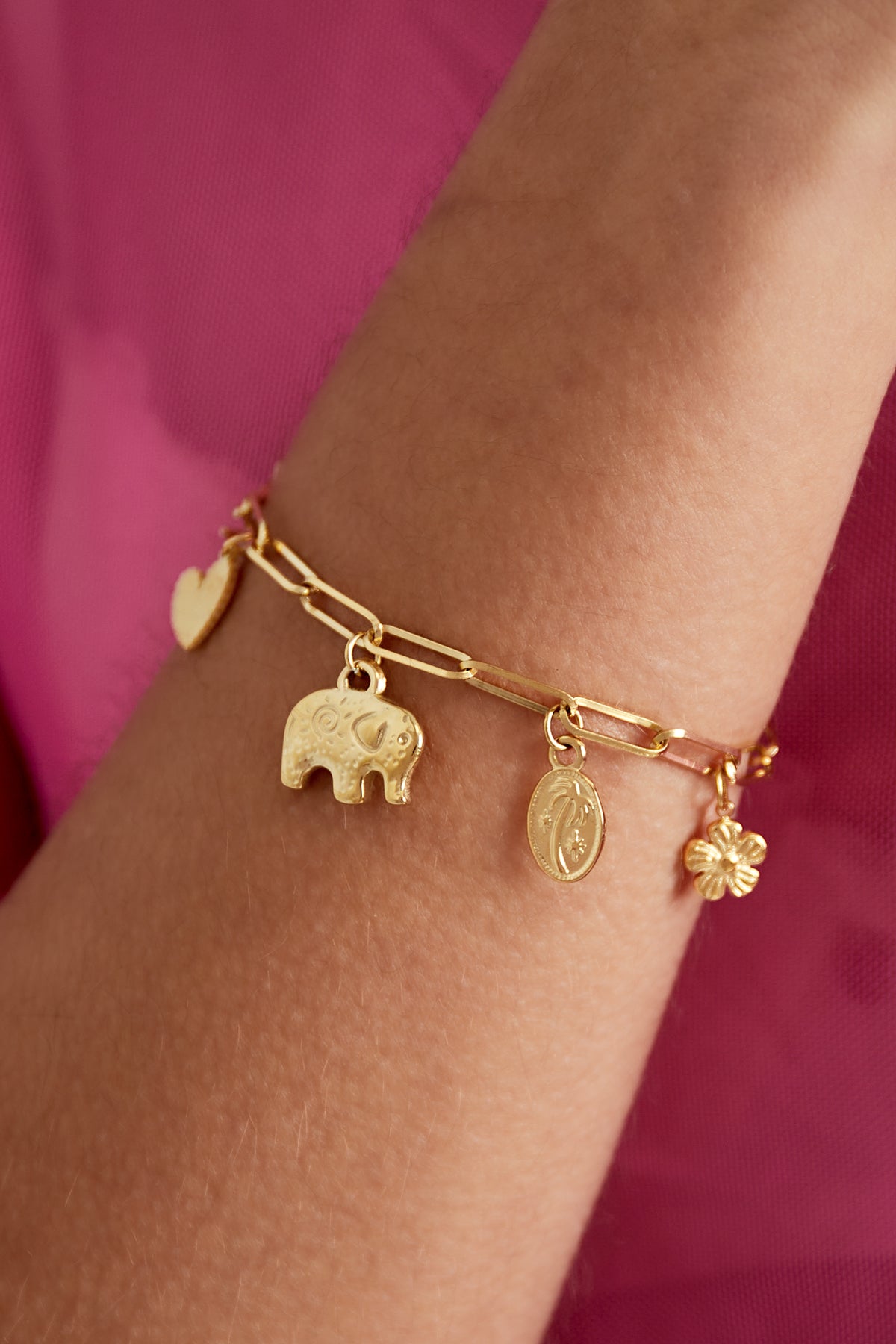 Close-up of golden charm link bracelet worn on wrist showing detailed elephant and flower charms against a pink background