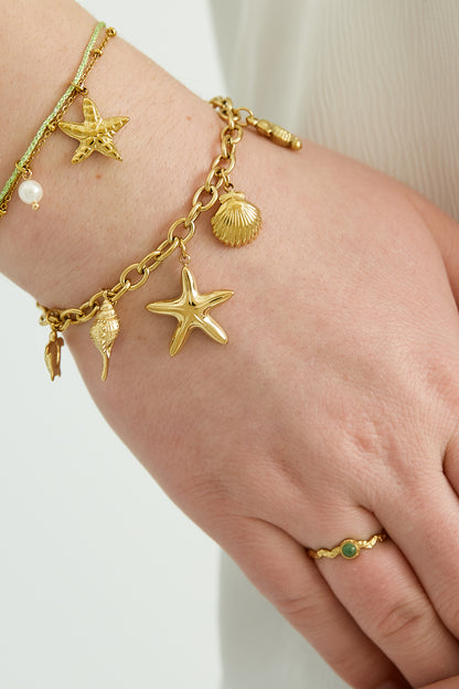 Gold sea charm bracelet worn on wrist alongside other delicate bracelets and a green gemstone ring, highlighting the shiny beach-themed charms