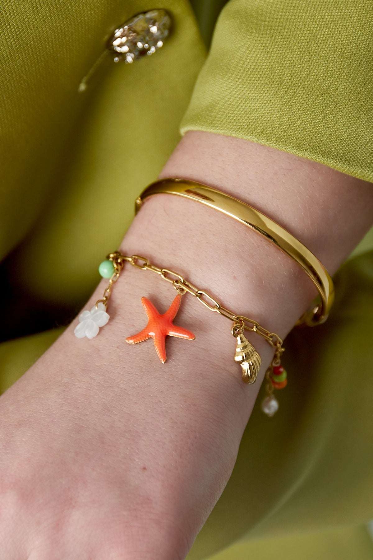 Woman's wrist wearing the gold-tone charm bracelet alongside a smooth gold bangle, styled with a green outfit