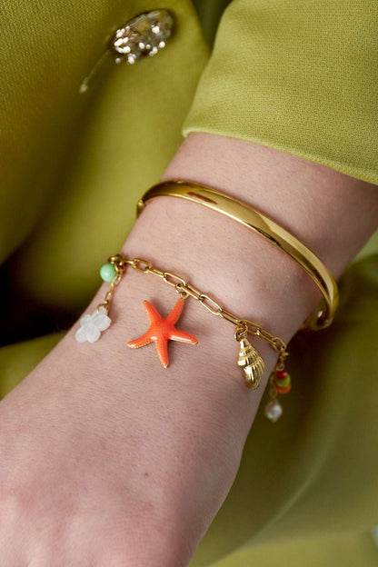 Woman's wrist wearing the gold-tone charm bracelet alongside a smooth gold bangle, styled with a green outfit