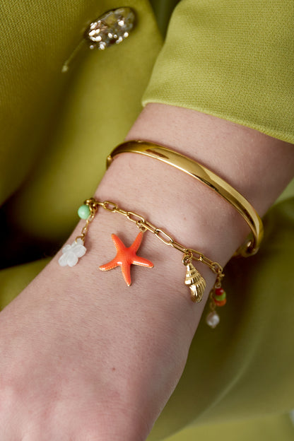 Woman's wrist wearing the gold-tone charm bracelet alongside a smooth gold bangle, styled with a green outfit