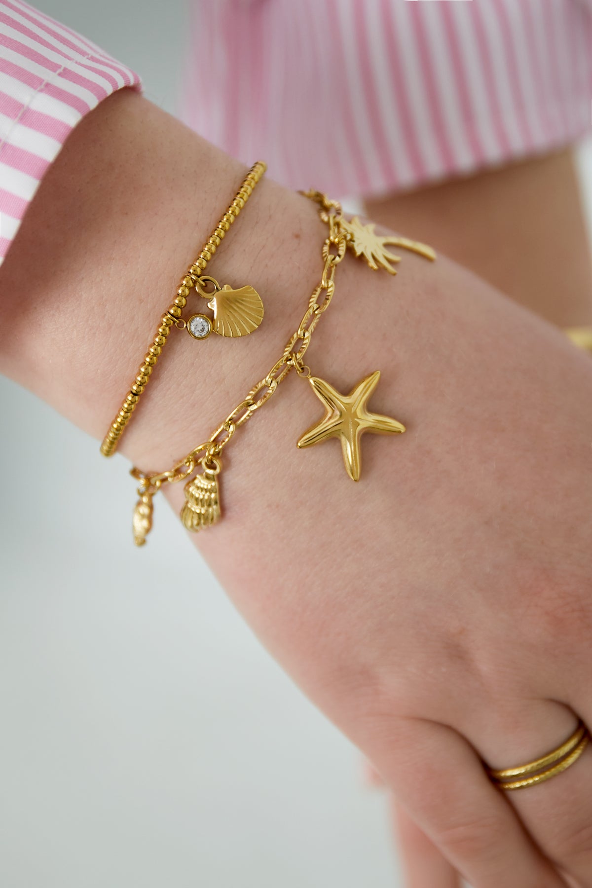 Wrist wearing gold charm bracelet with beach icons alongside a beaded bracelet on a woman’s hand in pink striped shirt