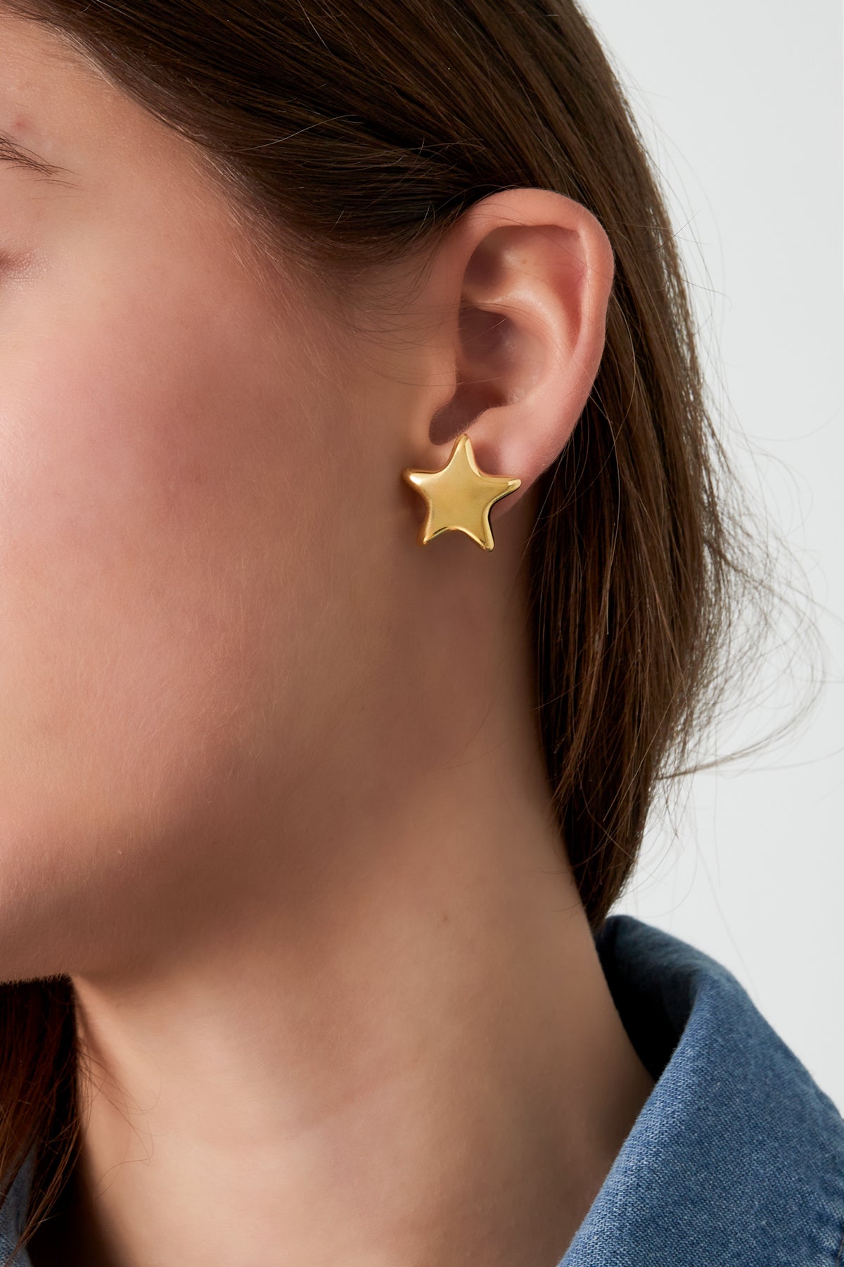 Model wearing a single gold star-shaped stud earring, styled with dark hair and denim jacket