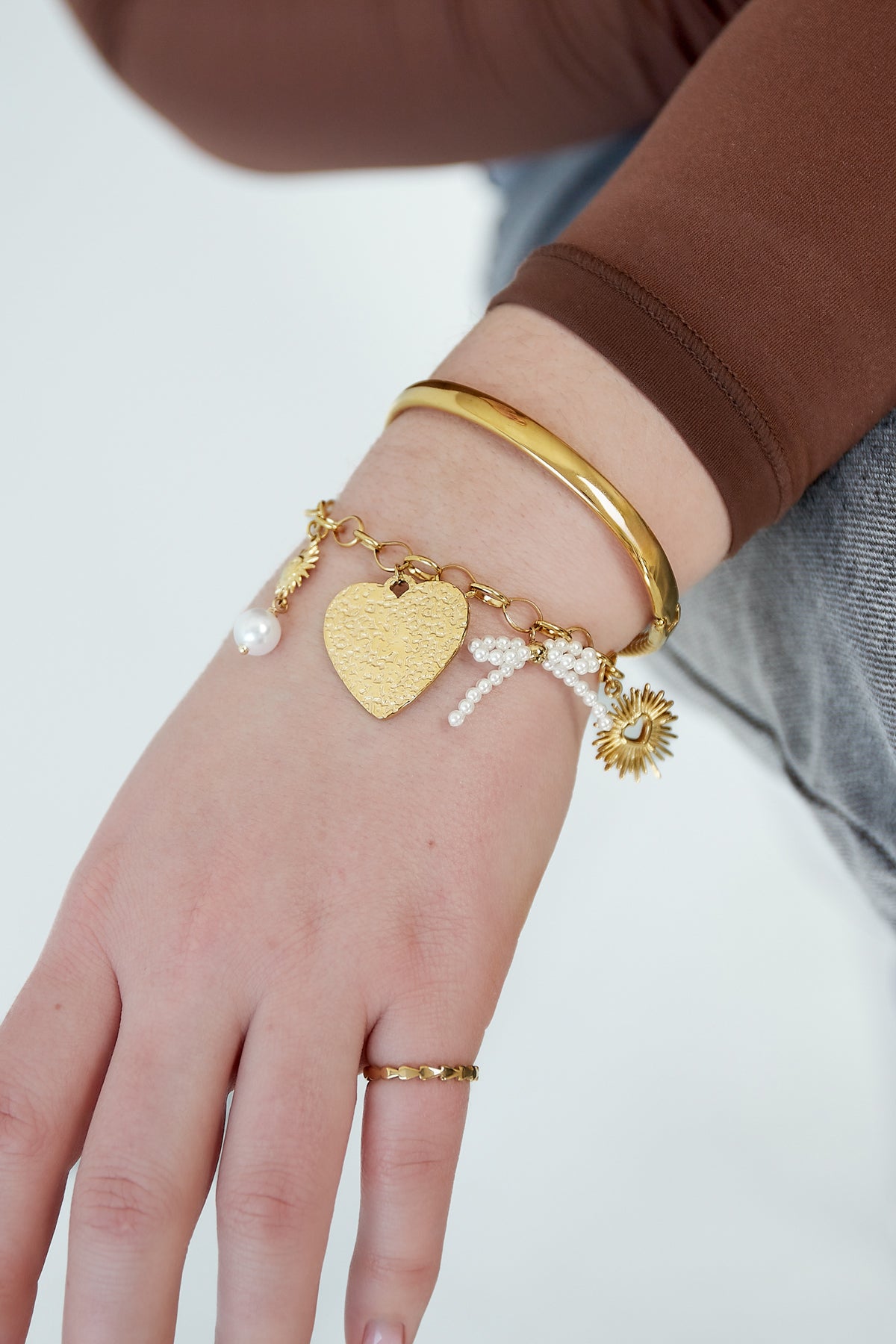 Close-up of wrist wearing gold-tone charm bracelet with heart pendant, sun and pearl charms alongside gold bangle and ring