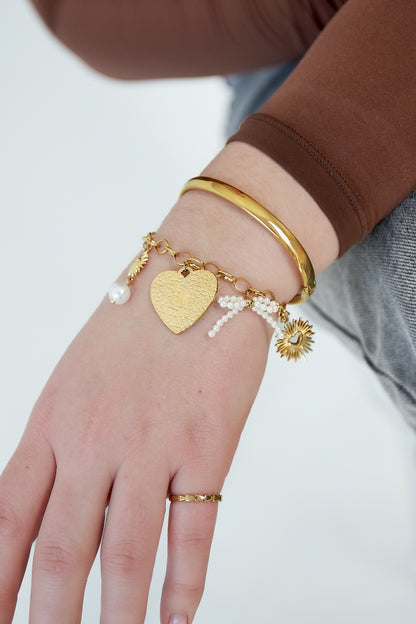 Close-up of wrist wearing gold-tone charm bracelet with heart pendant, sun and pearl charms alongside gold bangle and ring