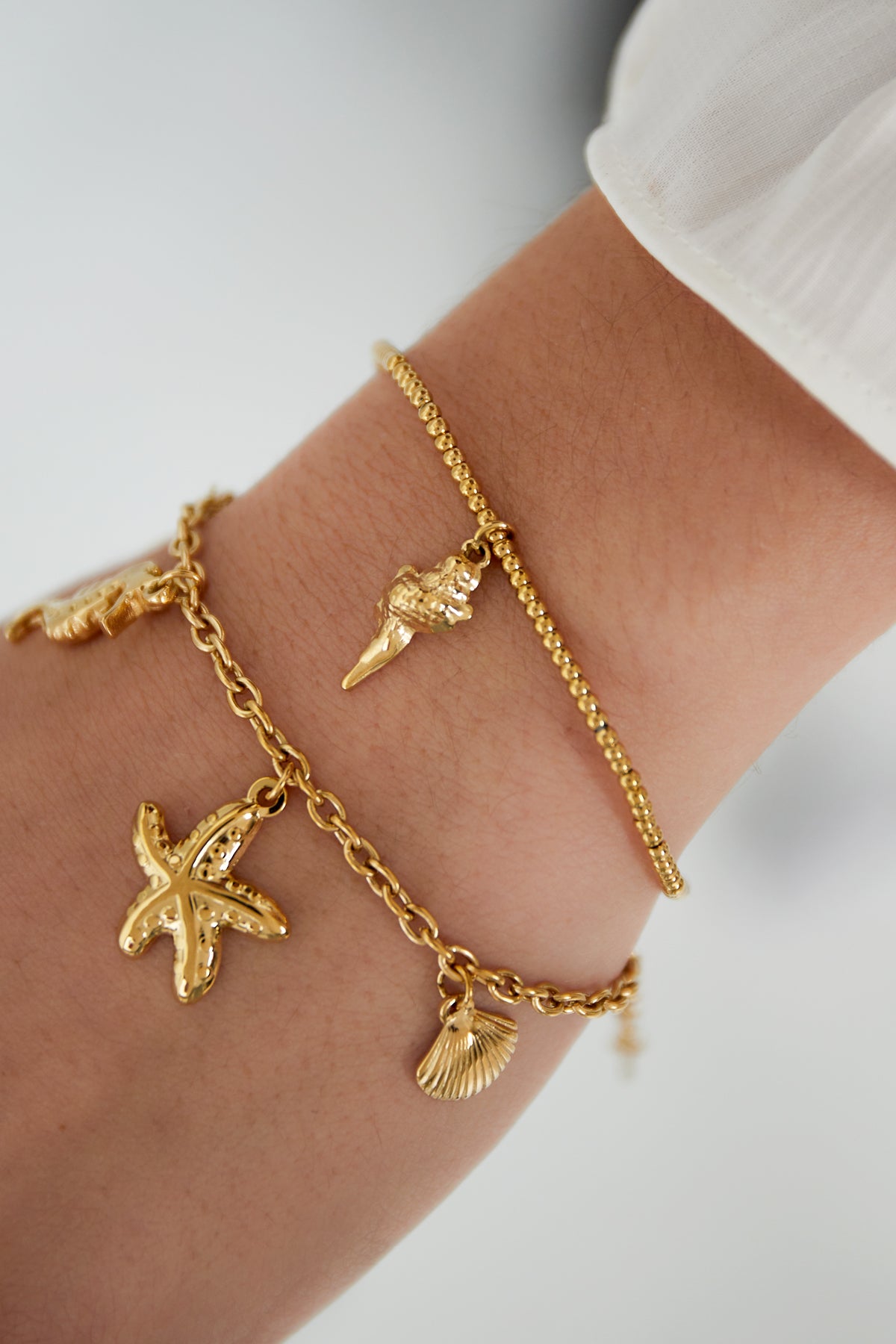 Woman's wrist adorned with two gold-tone bracelets, one beaded with a seashell charm and one chain with various charms
