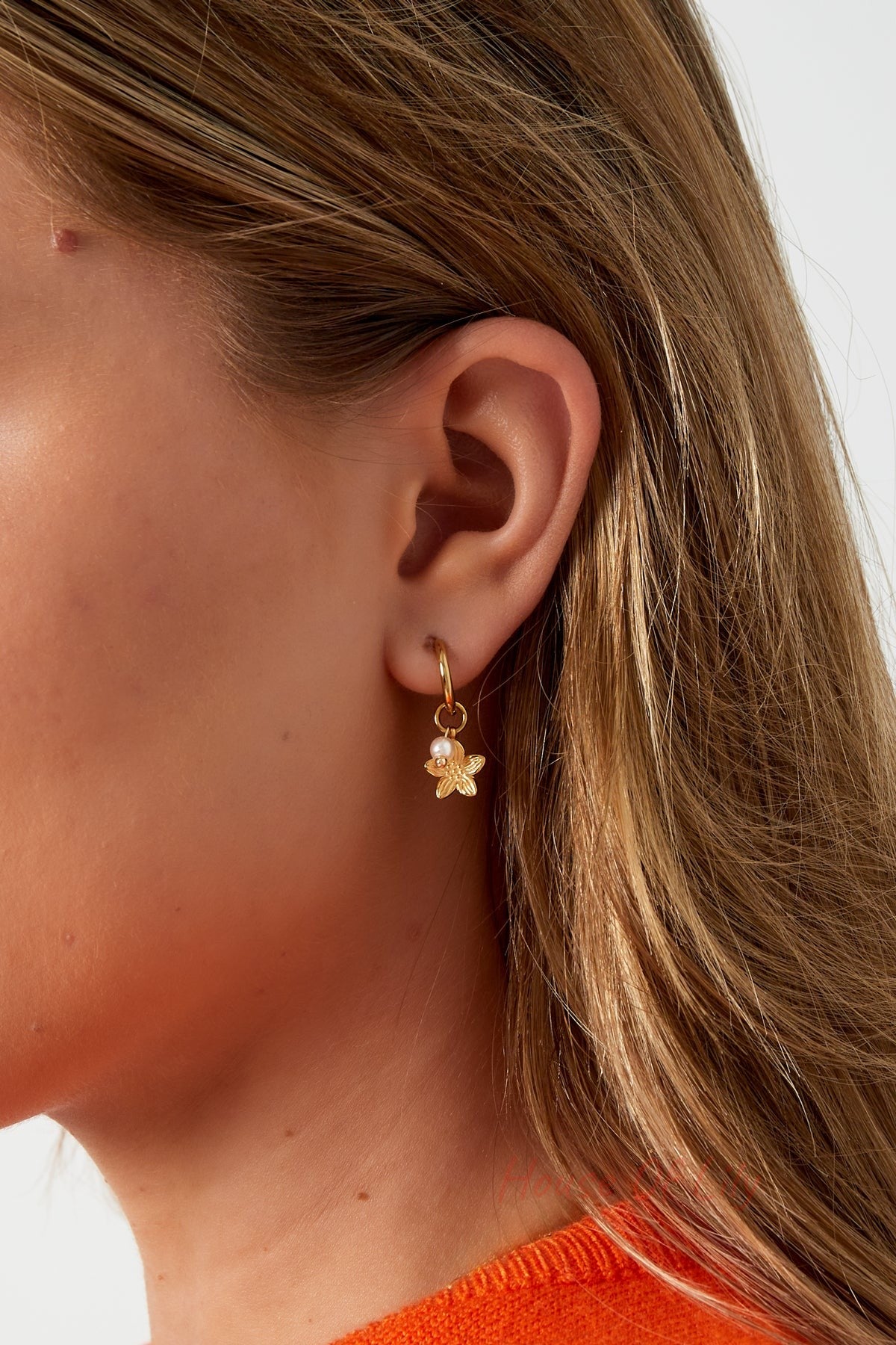 Close-up of gold floral charm hoop earring worn on a woman's ear with blonde hair and orange sweater
